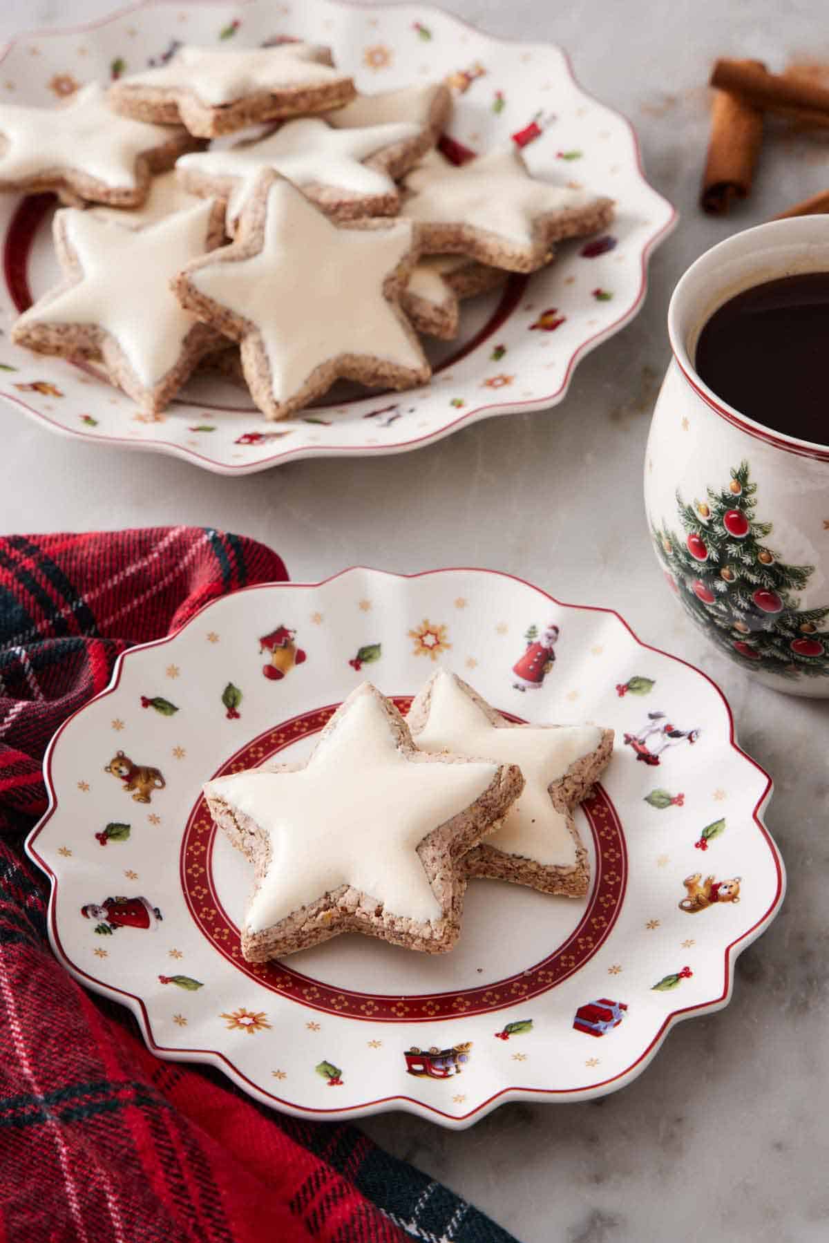 A plate with two star shaped zimtsterne with more in the background on a larger platter. A coffee beside it.