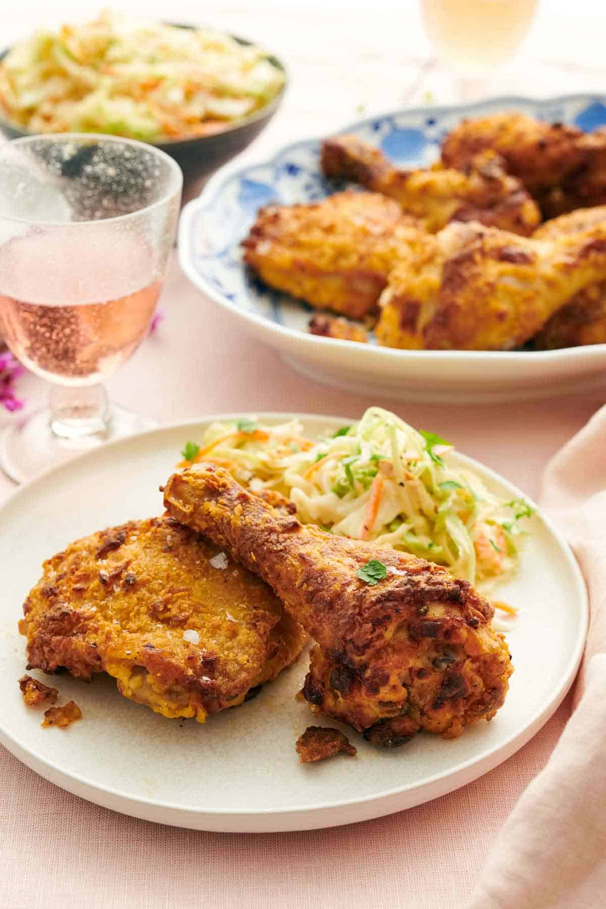 A plate with two pieces of air fryer fried chicken along with coleslaw. A drink and platter of chicken in the background.