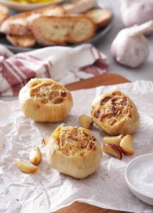 Three air fryer roasted garlic also with roasted cloves on a parchment-lined wooden serving board. More garlic and sliced bread in the background.