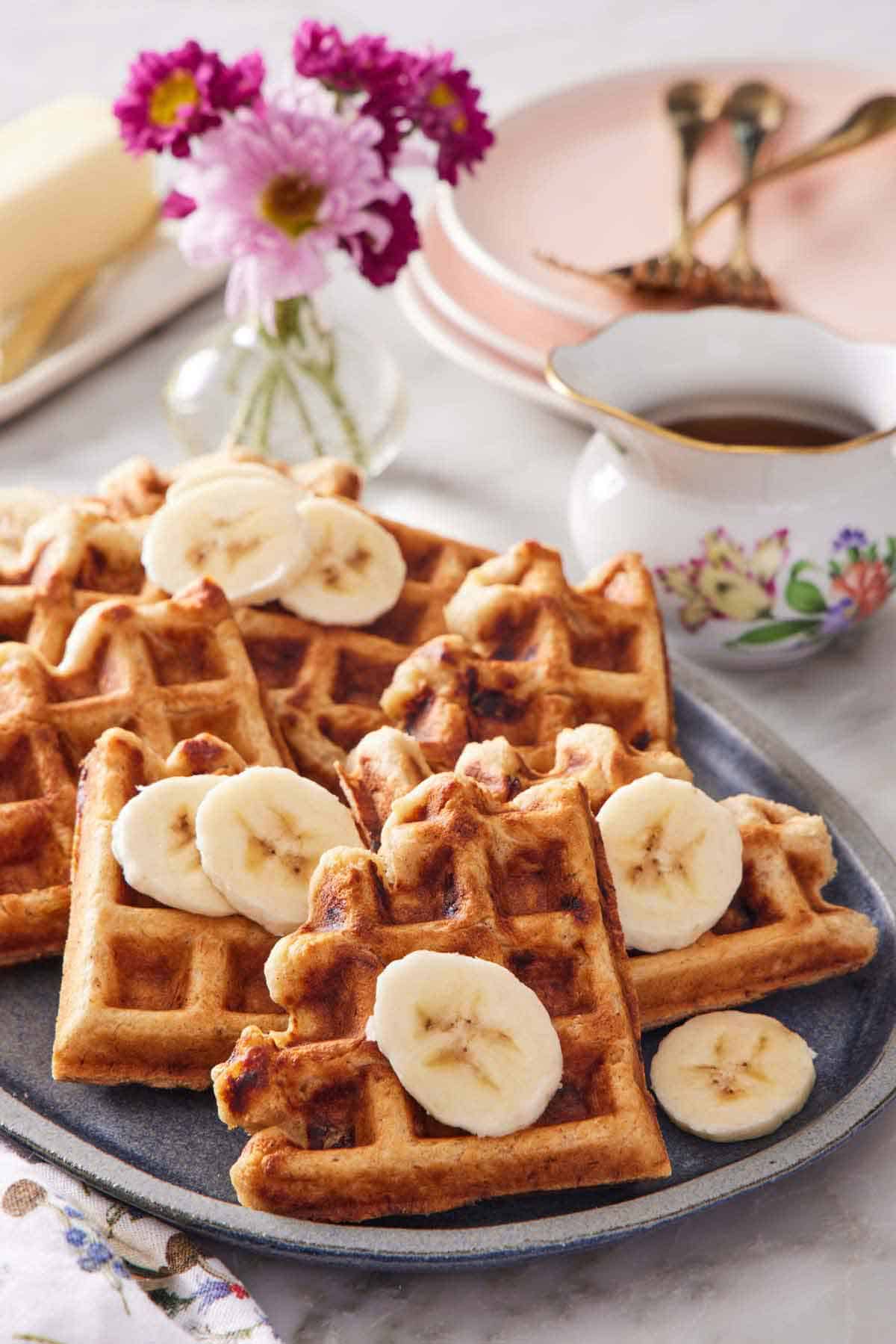 A platter with pieces of banana waffles topped with sliced bananas. Syrup in the background along with flowers, stacks of plates. and forks.