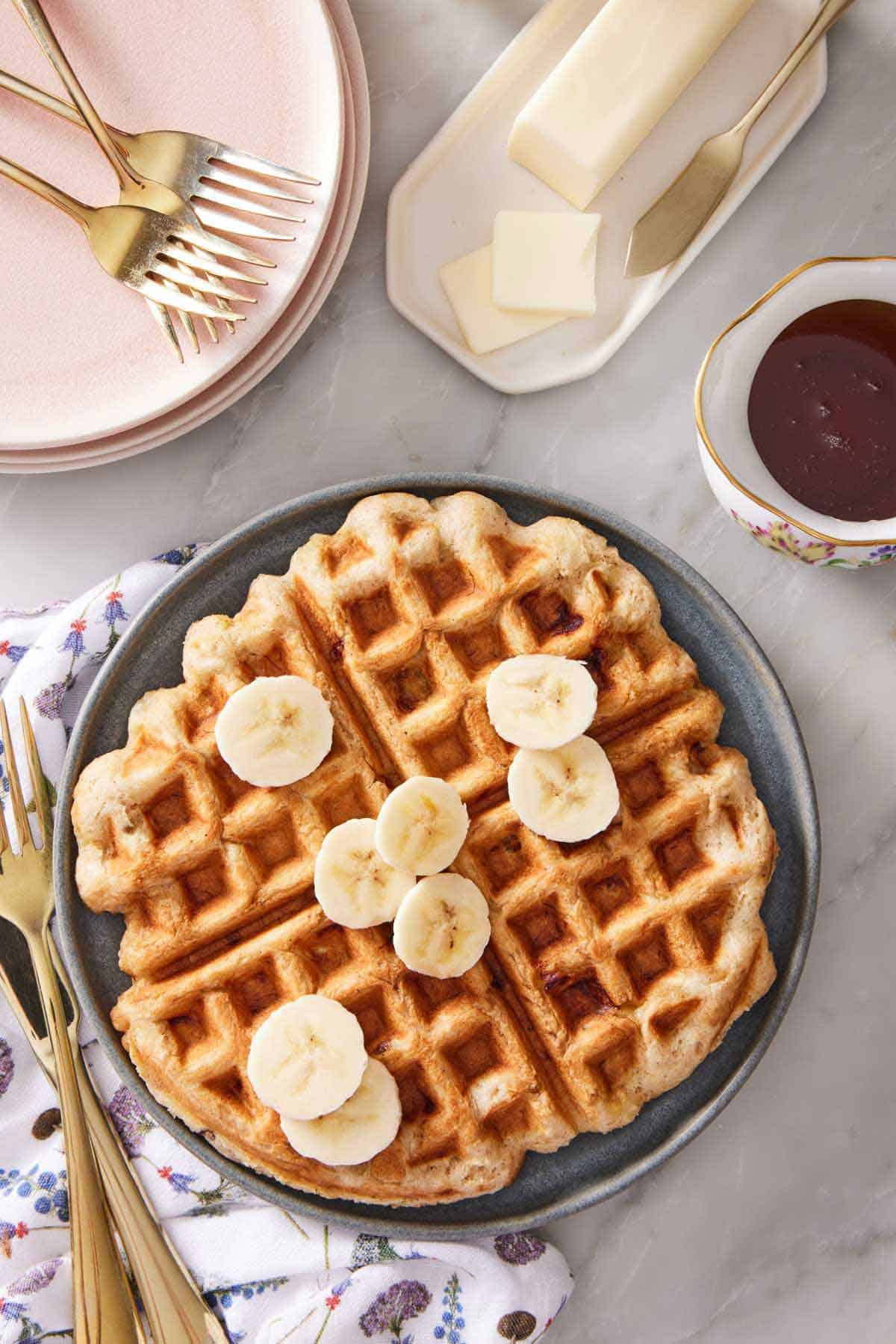 Overhead view of a plate of banana waffle topped with sliced bananas. A stack of plates, forks, syrup, and butter off to the side.