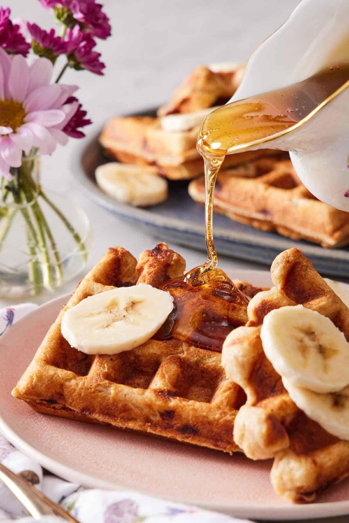 Syrup being poured onto a piece of banana waffle with banana slices. Flowers and more waffles in the background.