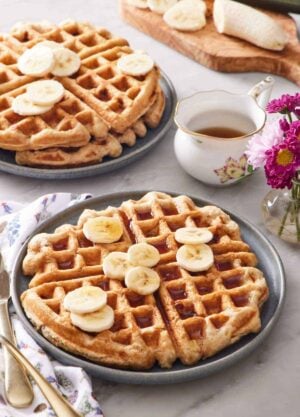 A plate with a large banana waffle topped with syrup and sliced bananas. A stack of two waffles in the back along with syrup and a banana being sliced.