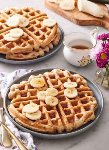 A plate with a large banana waffle topped with syrup and sliced bananas. A stack of two waffles in the back along with syrup and a banana being sliced.