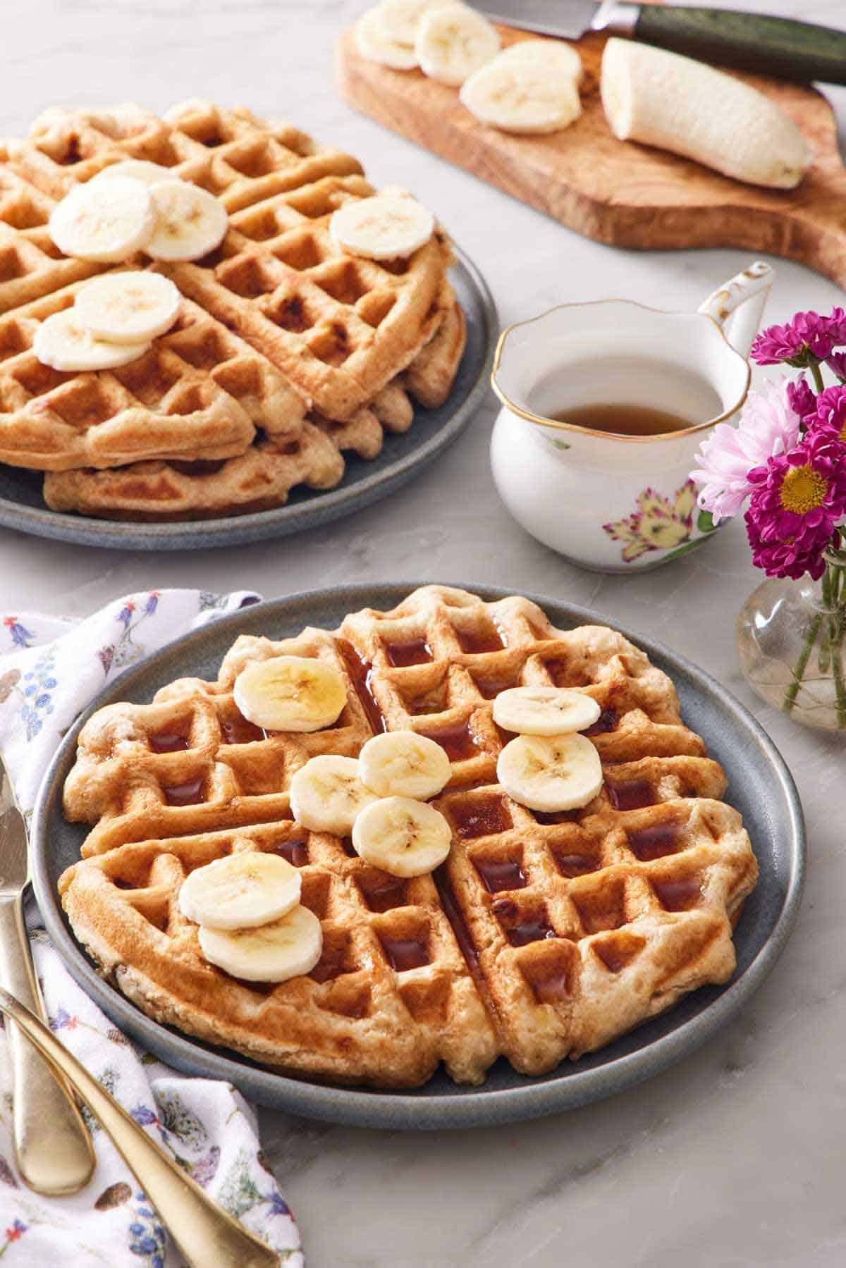 A plate with a large banana waffle topped with syrup and sliced bananas. A stack of two waffles in the back along with syrup and a banana being sliced.