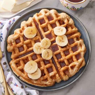 Overhead view of banana waffles on a plate with sliced bananas and syrup. Forks, plates, linen, flowers, and butter off to the side.