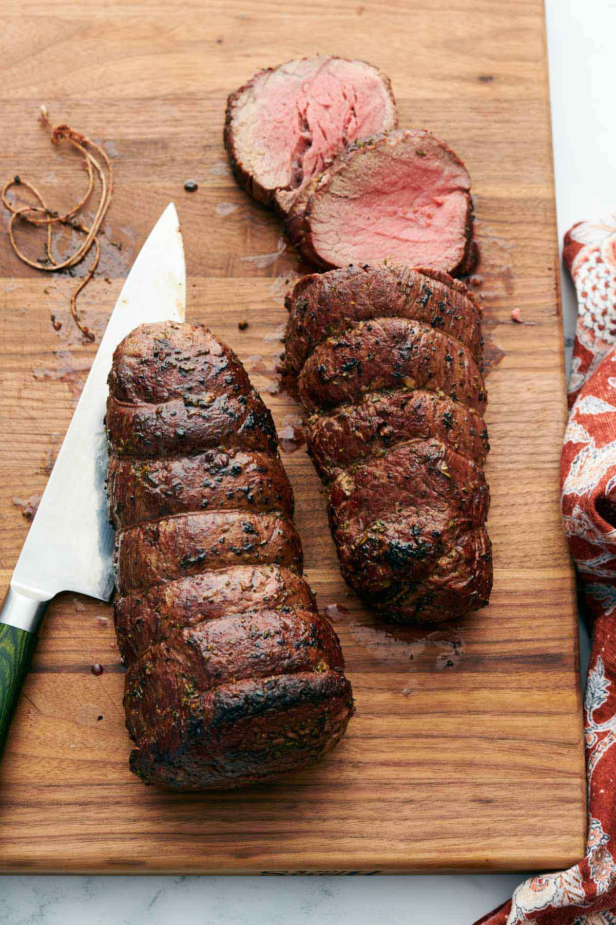 Overhead view of a wooden cutting board with beef tenderloin in the process of being cut. A knife on the side.