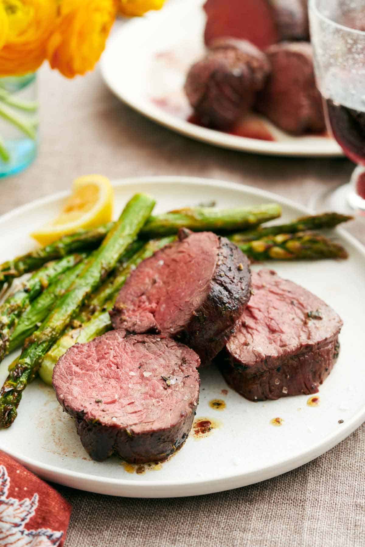 A plate with three pieces of beef tenderloin with roasted asparagus along with a lemon wedge. Flowers, glass of wine, and more beef tenderloin in the background.