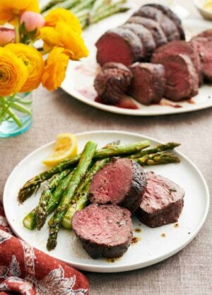 A plate with beef tenderloins along with asparagus and a lemon wedge. A platter with more beef tenderloin in the background along with flowers in a vase.