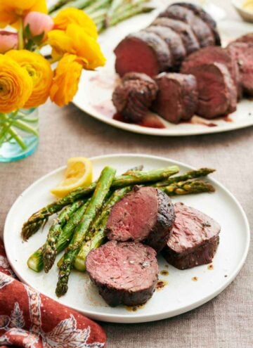 A plate with beef tenderloins along with asparagus and a lemon wedge. A platter with more beef tenderloin in the background along with flowers in a vase.