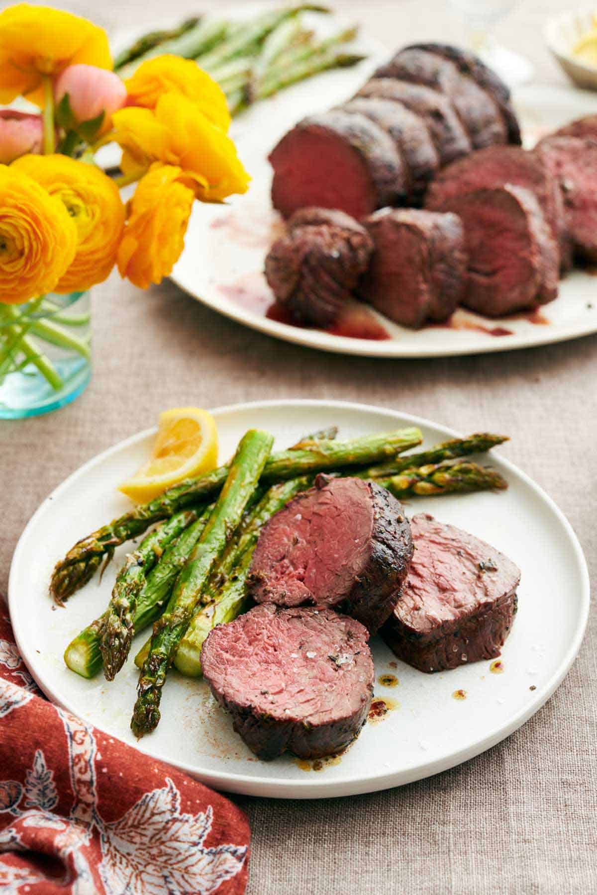 A plate with beef tenderloins along with asparagus and a lemon wedge. A platter with more beef tenderloin in the background along with flowers in a vase.