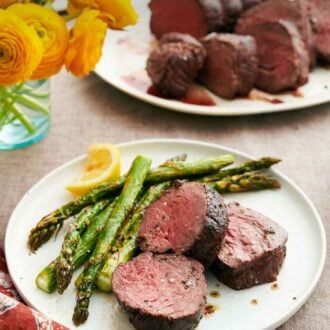 Pinterest graphic of a plate with beef tenderloins along with asparagus and a lemon wedge. A platter with more beef tenderloin in the background.