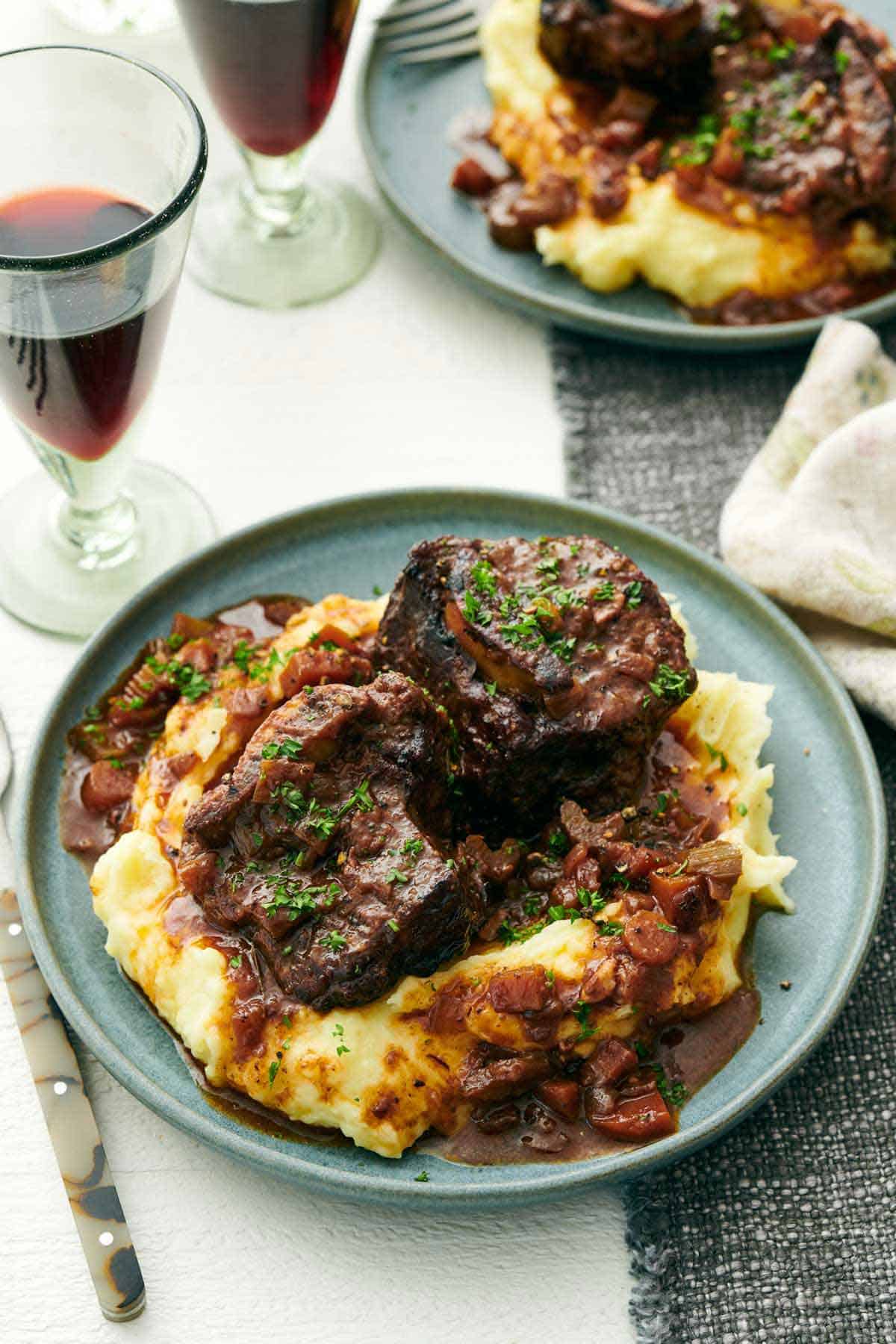 A plate of braised beef short ribs over mashed potatoes. Two glasses of wine and another plate of braised beef short ribs in the background.