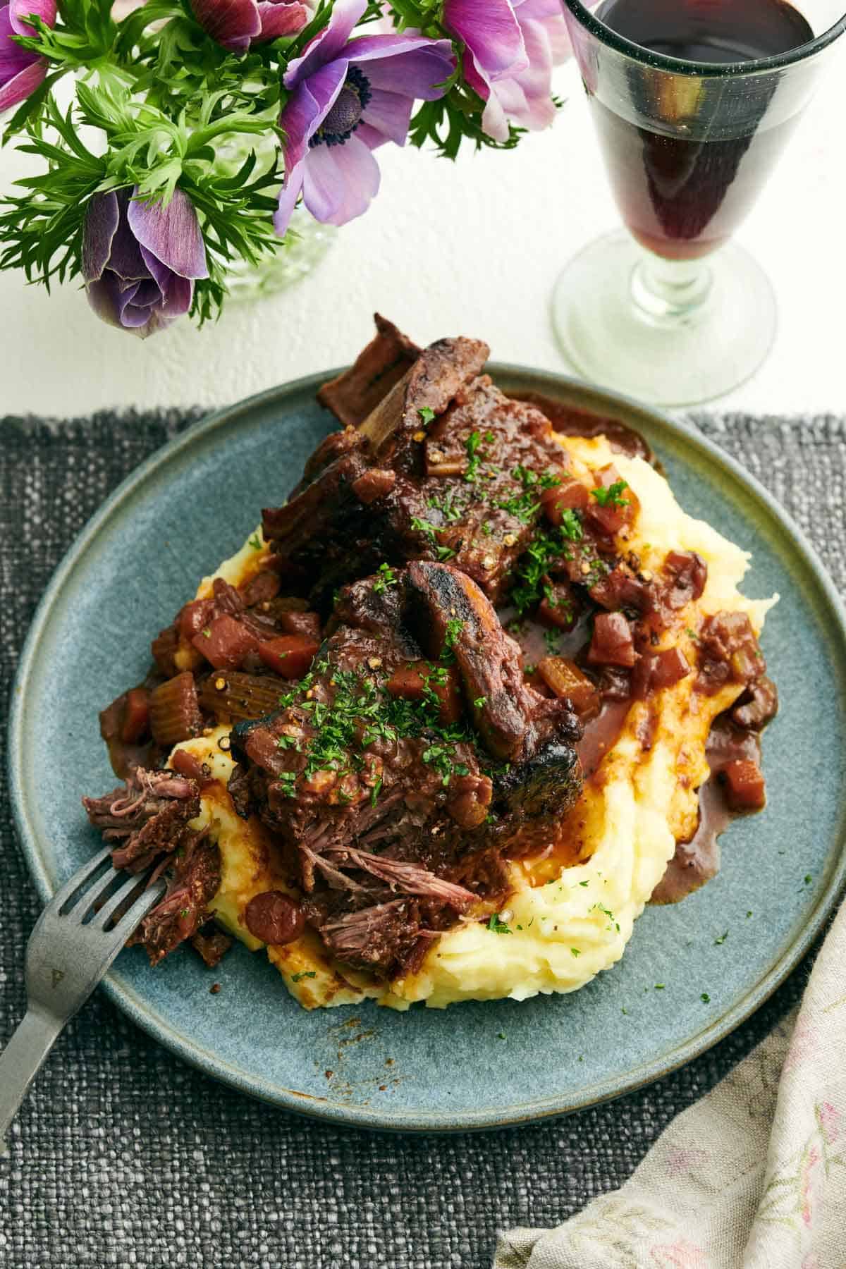 A plate of braised beef short ribs over mashed potatoes. A drink and some flowers off to the side.