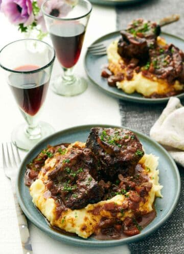 A plate of mashed potatoes with braised beef short ribs on top. A second plate and two glasses of wine in the background.