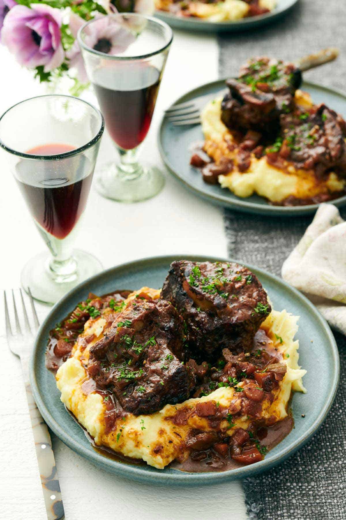 A plate of mashed potatoes with braised beef short ribs on top. A second plate and two glasses of wine in the background.