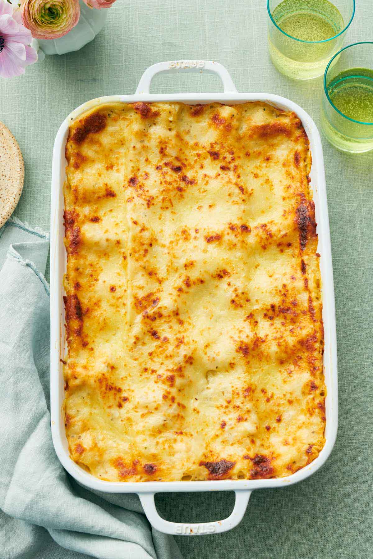 Overhead view of a baking dish of chicken lasagna with drinks on the side along with some flowers.