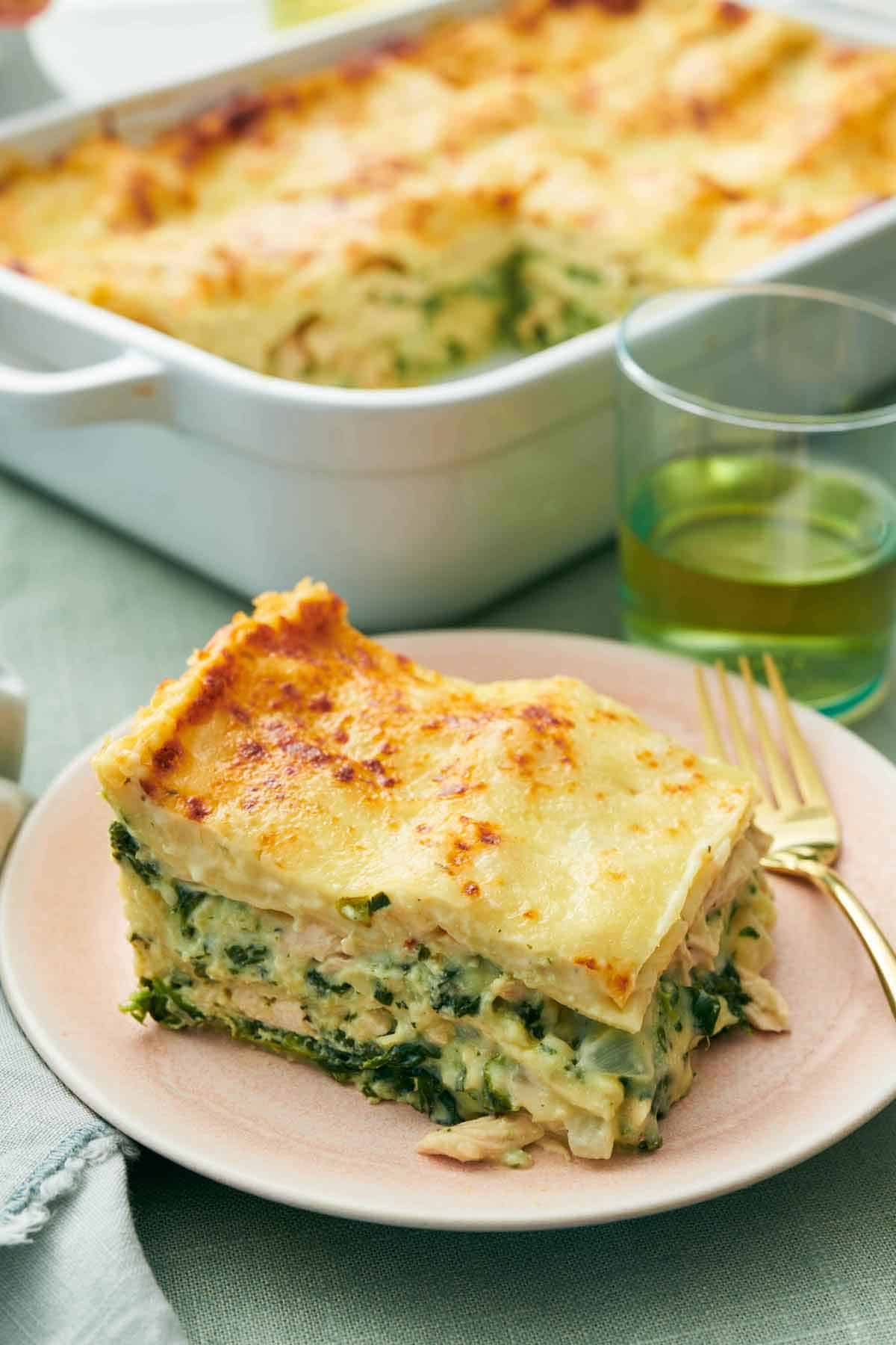 A slice of chicken lasagna on a pink plate with a fork. Baking dish with the rest of the chicken lasagna in the background.