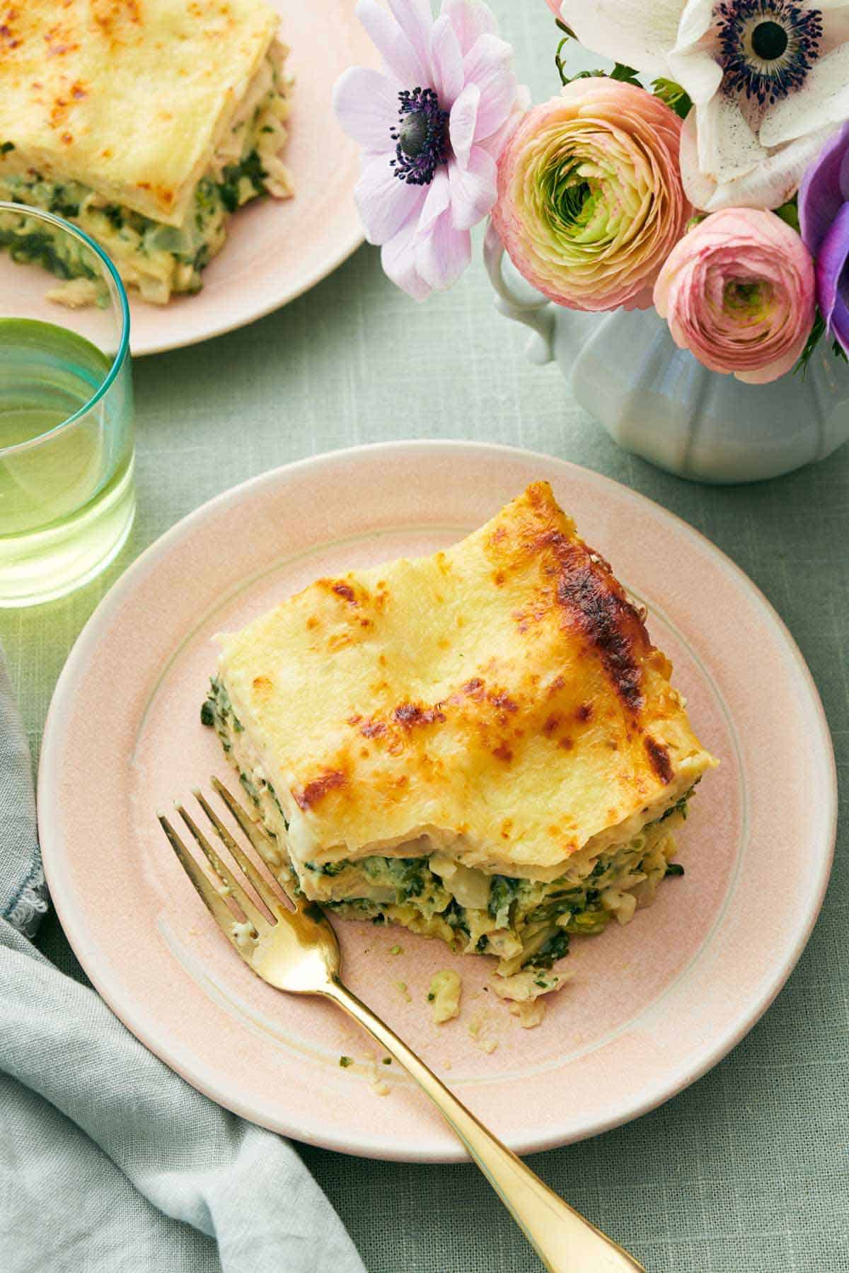 Overhead view of a slice of chicken lasagna with a bite taken out. A fork beside the lasagna slice and flowers, a drink, and another plated slice off to the side.