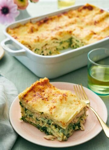 A plate with a slice of chicken lasagna with a fork. A baking dish with the rest of the lasagna in the background with a drink and some flowers.