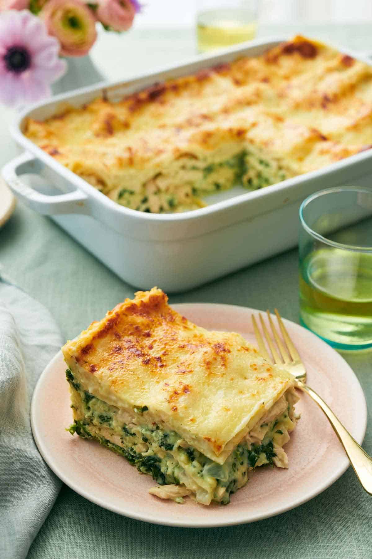 A plate with a slice of chicken lasagna with a fork. A baking dish with the rest of the lasagna in the background with a drink and some flowers.