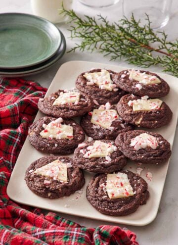 A platter of chocolate peppermint bark cookies. Decorative pines, plates, and a checkered linen napkin surrounding the platter.