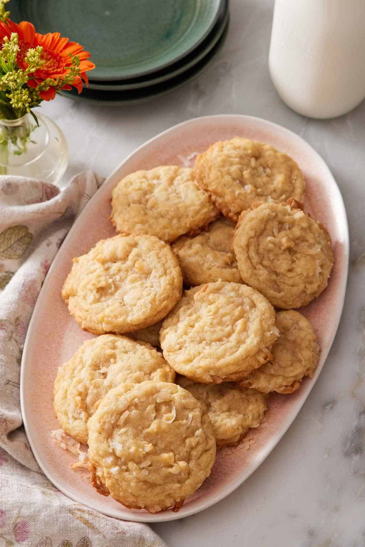 A pink platter with multiple coconut cookies. Flowers, a stack of plates, and milk in the background.