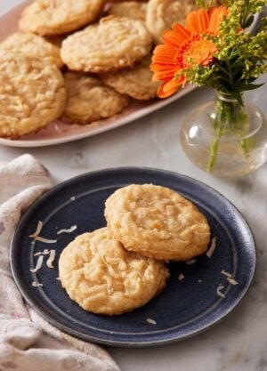 A plate with two coconut cookies. A vase with flowers and more cookies on a platter in the background.