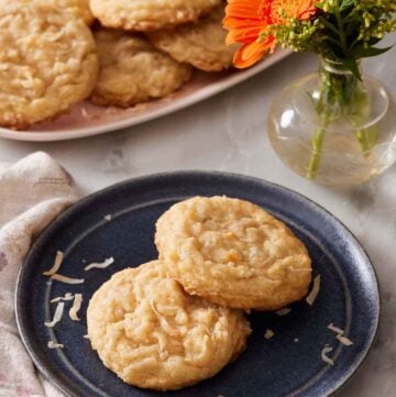 A plate with two coconut cookies. A vase with flowers and more cookies on a platter in the background.