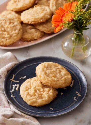 A plate with two coconut cookies. A vase with flowers and more cookies on a platter in the background.