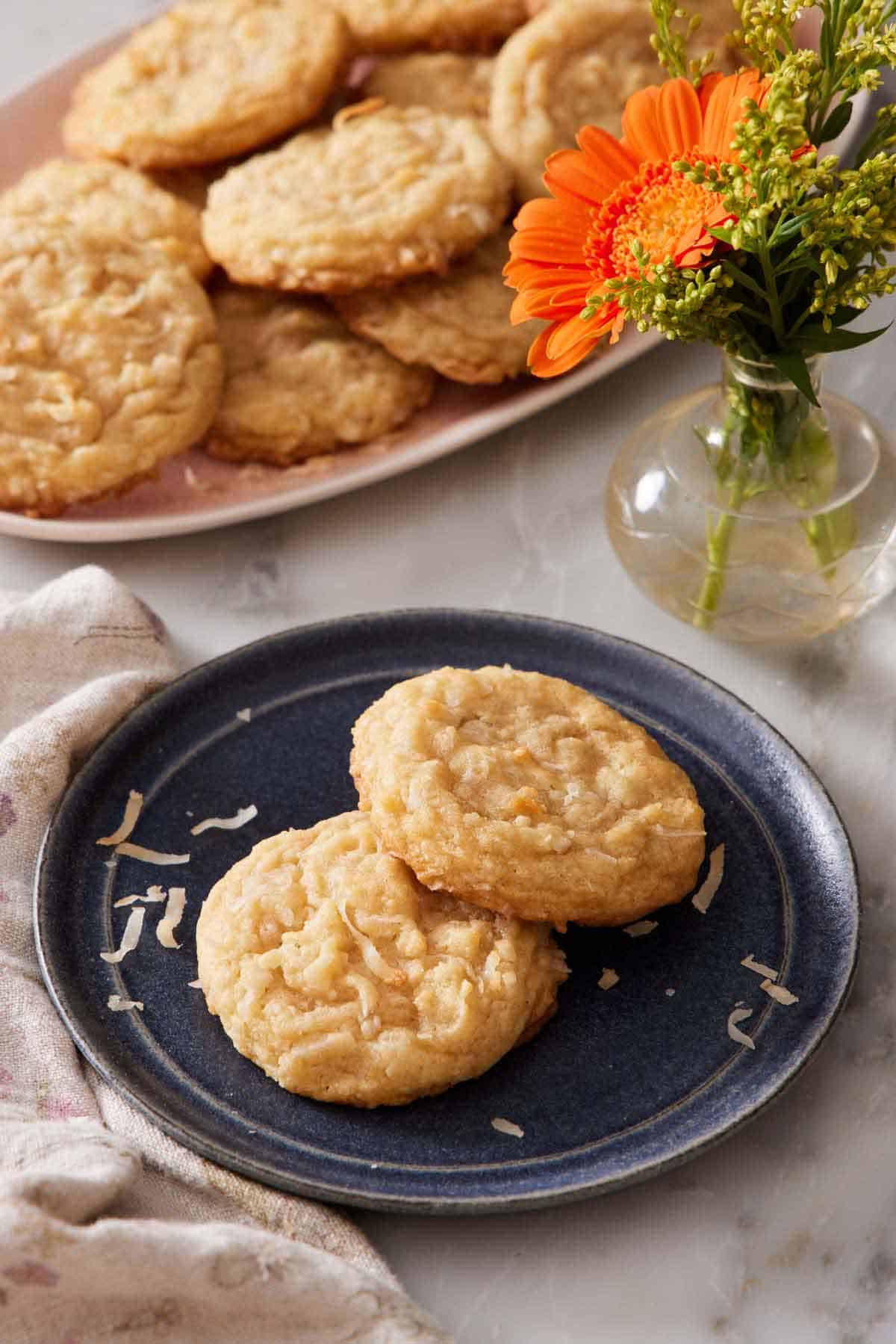 A plate with two coconut cookies. A vase with flowers and more cookies on a platter in the background.