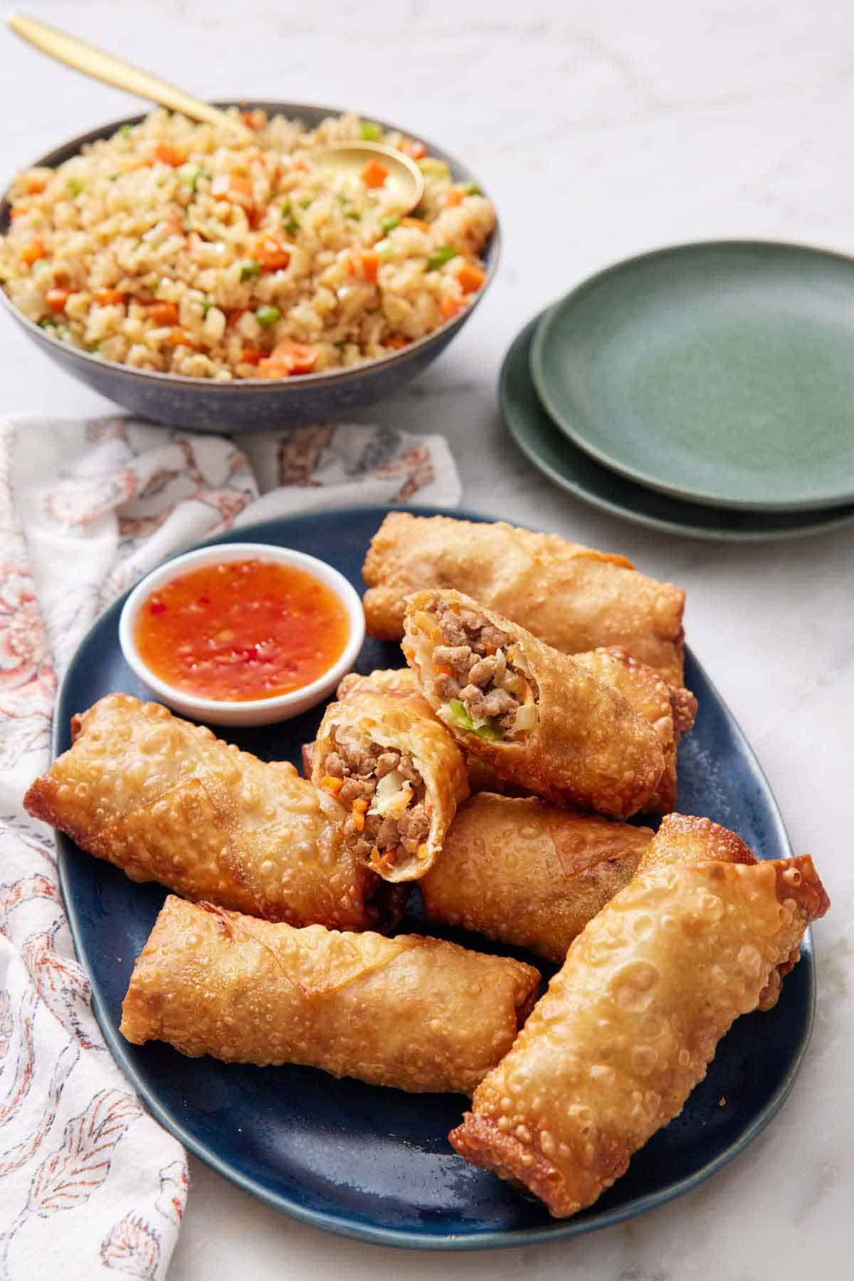 A platter of egg rolls with one cut in half on top. A small bowl of dip in the platter and fried rice in the background along with two plates.