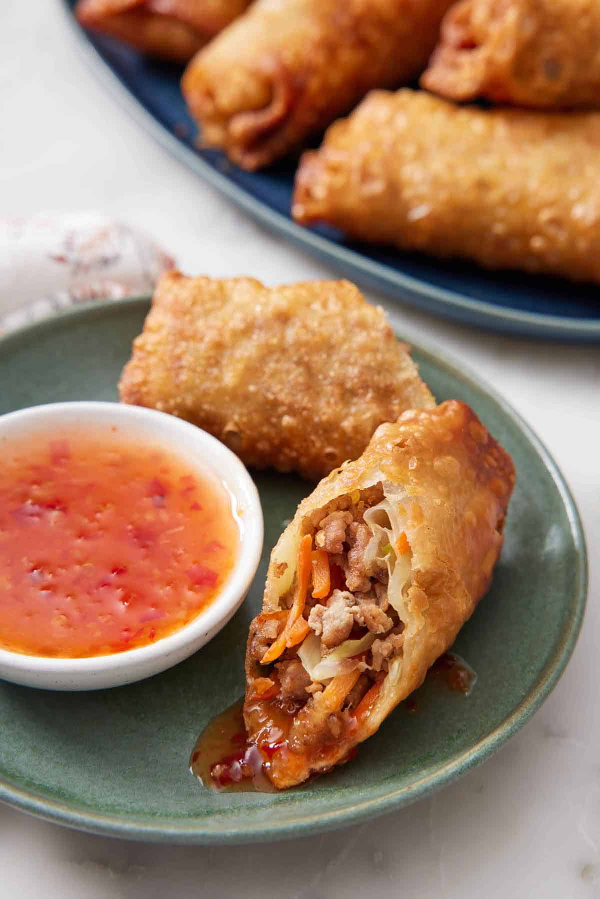 A plate with an egg roll cut in half with some sauce on it with the bowl of sauce on the side.