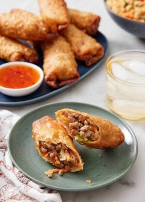 A plate with an egg rolls cut in half on a plate. A drink and platter of egg rolls with dip in the background.