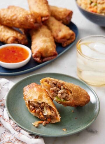 A plate with an egg rolls cut in half on a plate. A drink and platter of egg rolls with dip in the background.