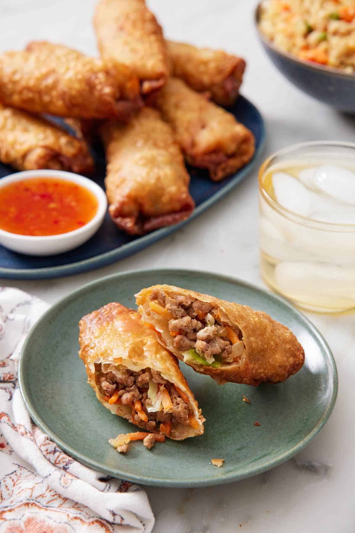 A plate with an egg rolls cut in half on a plate. A drink and platter of egg rolls with dip in the background.