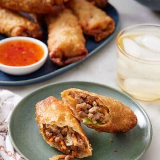 Pinterest graphic of a plate with an egg rolls cut in half on a plate. A drink and platter of egg rolls with dip in the background.