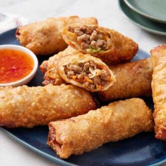 A platter of egg rolls with an opened one on top, showing the interior, with a bowl of sauce on the side.