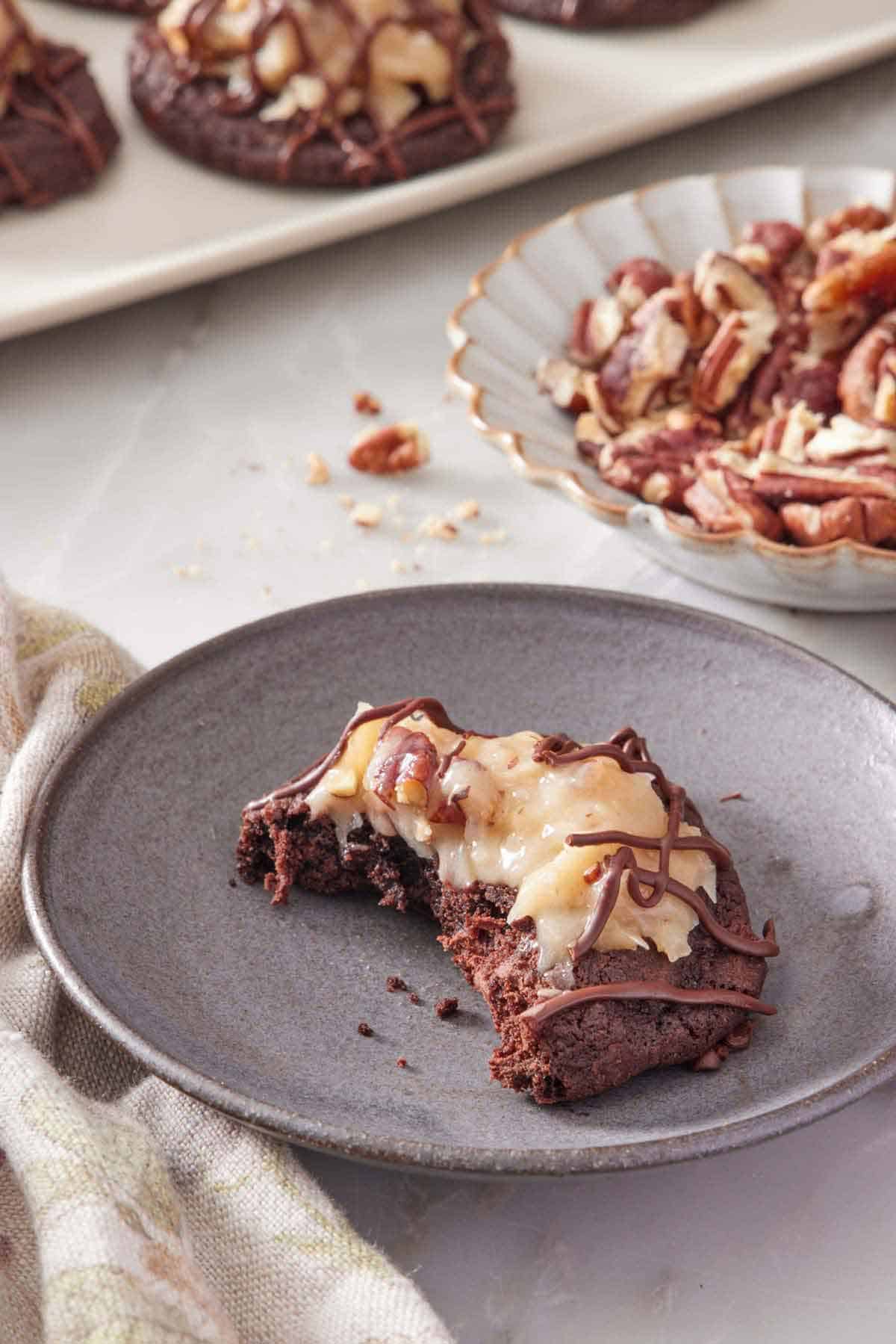 A plate with a German chocolate cookie with a bite taken out of it. Pecans in the background.