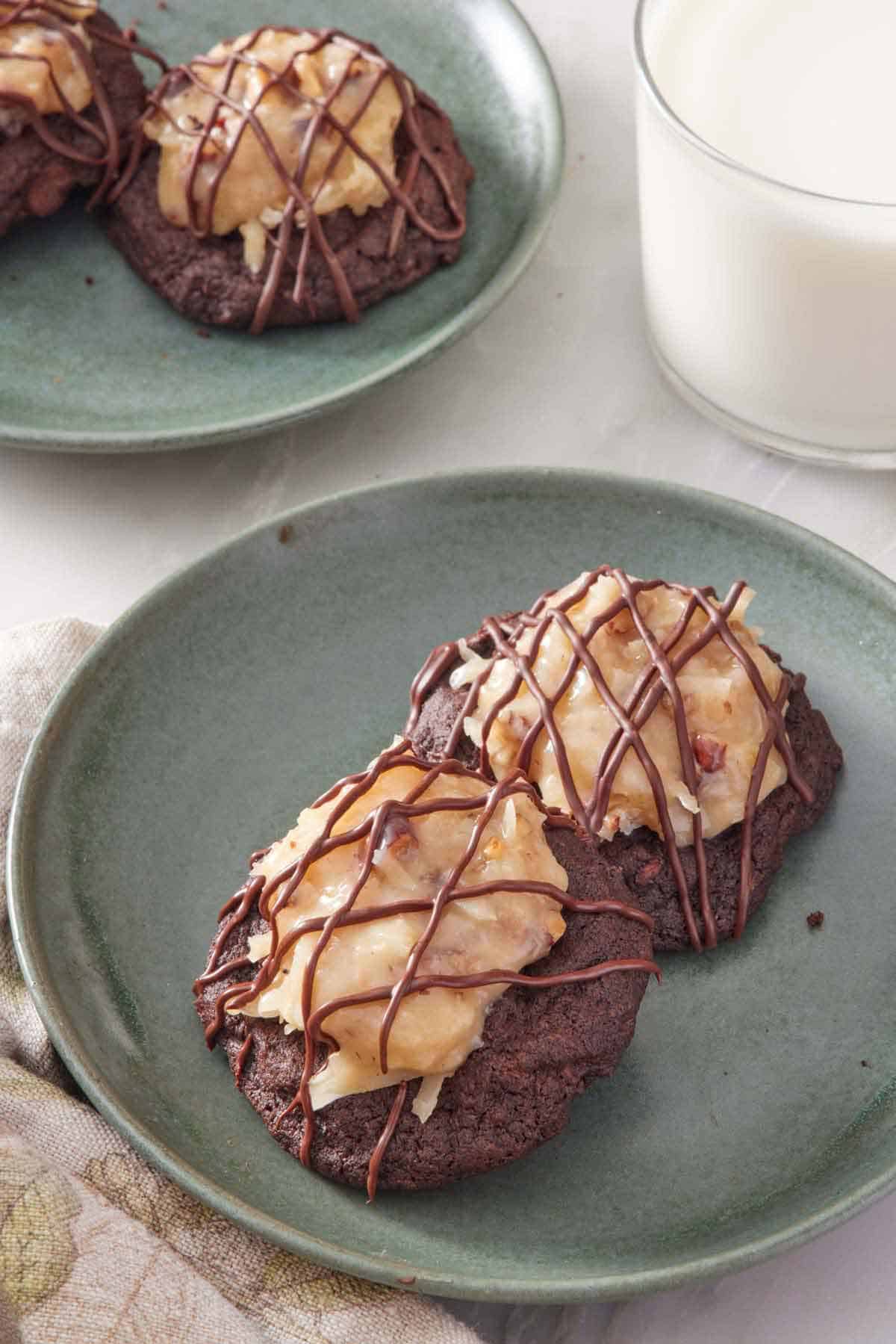 A plate with two German chocolate cookies. A glass of milk in the back along with more cookies.