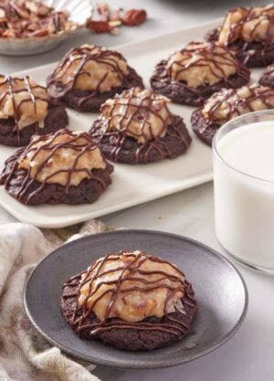 A plate with a German chocolate cookie. A platter with more cookies in the back along with a glass of milk.