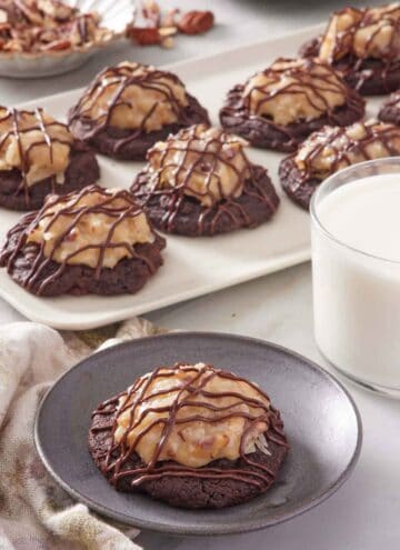 A plate with a German chocolate cookie. A platter with more cookies in the back along with a glass of milk.