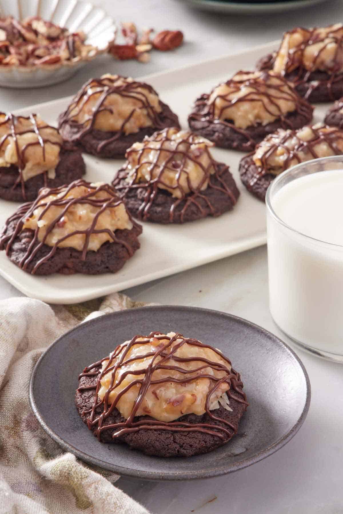 A plate with a German chocolate cookie. A platter with more cookies in the back along with a glass of milk.