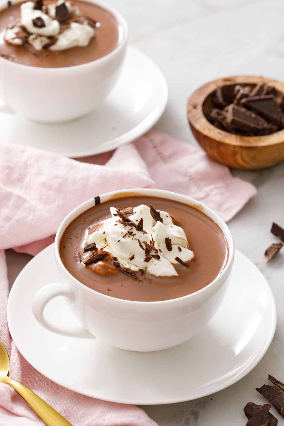 A mug of hot chocolate topped with whipped cream and shabed chocolate. Another mug and a bowl of chocolate in the background.