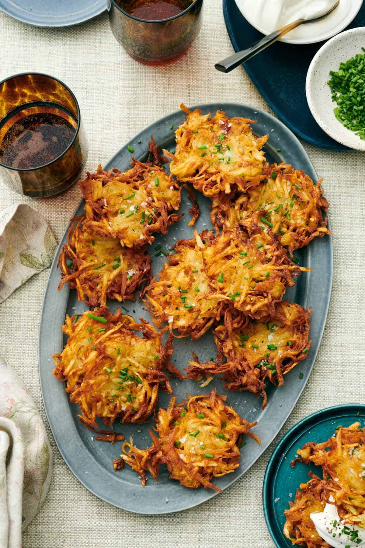 Overhead view of multiple crispy potato latkes on an oval platter. Drinks, chives, sour cream, and more latkes on the side.