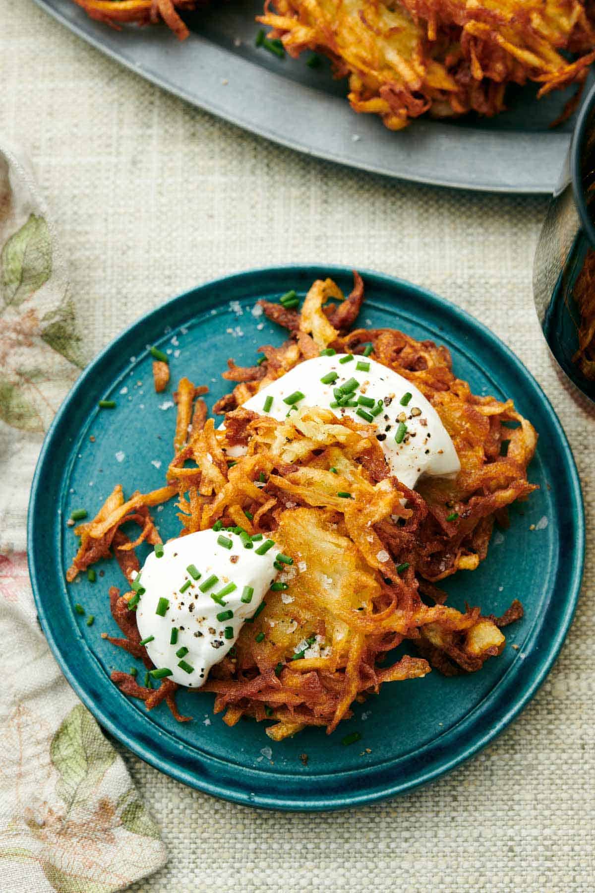 Overhead view of a blue plate with two potato latkes topped with sour cream, chives, and pepper.