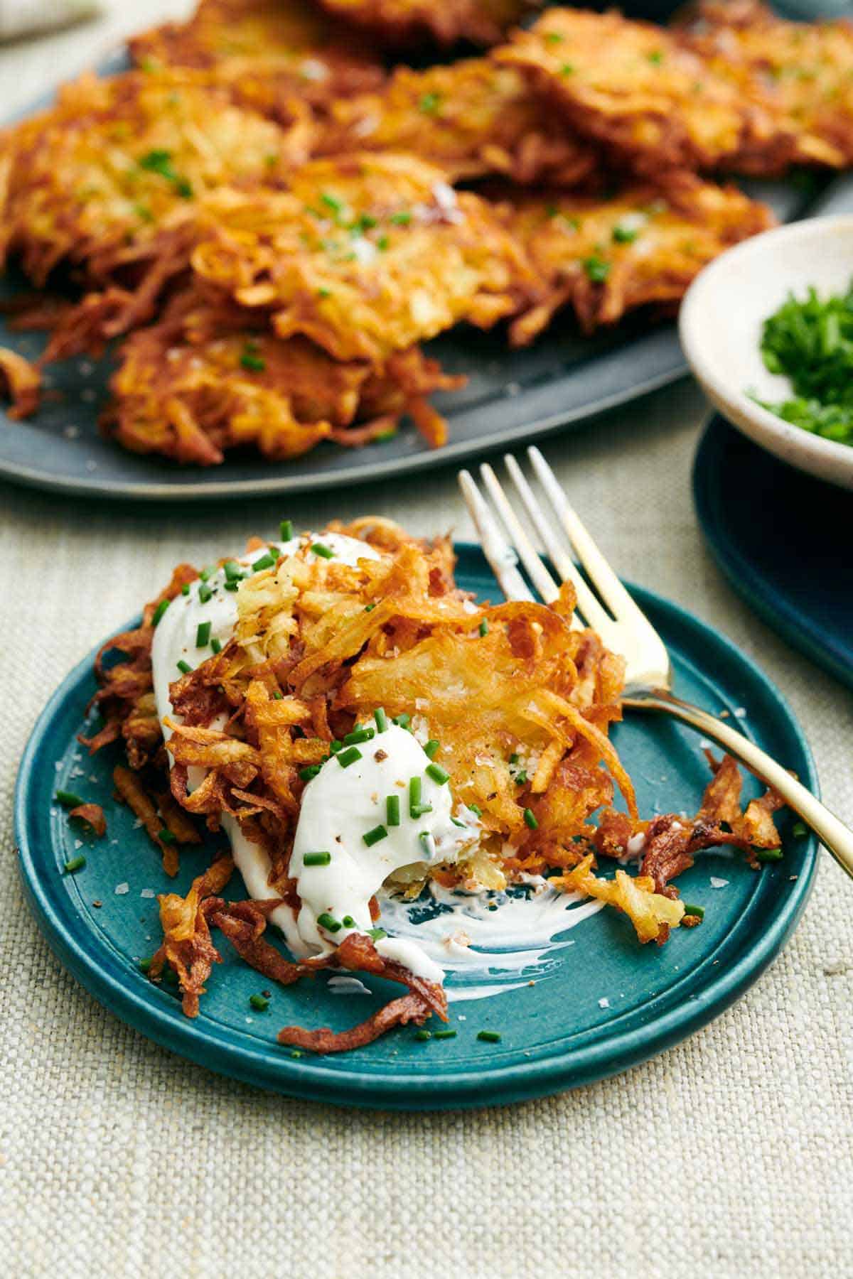 A plate with potato latkes, partially eaten, with a fork. A platter of latkes in the background with some chives.