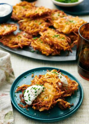A plate with two potato latkes topped with sour cream and chives. More on a platter in the background with a drink.