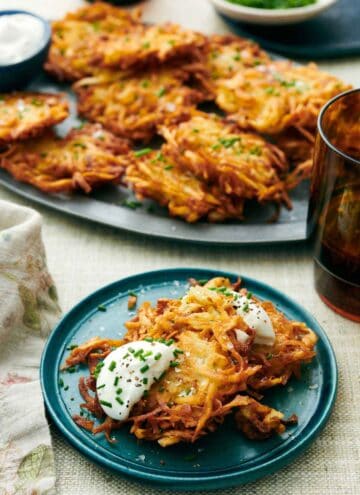 A plate with two potato latkes topped with sour cream and chives. More on a platter in the background with a drink.