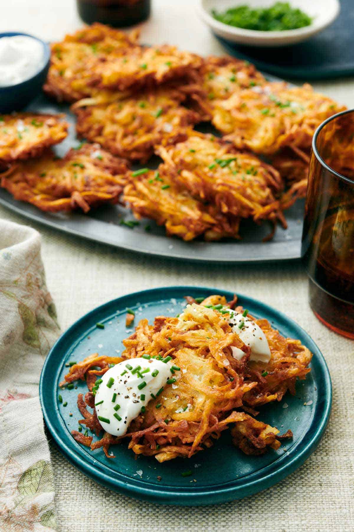 A plate with two potato latkes topped with sour cream and chives. More on a platter in the background with a drink.